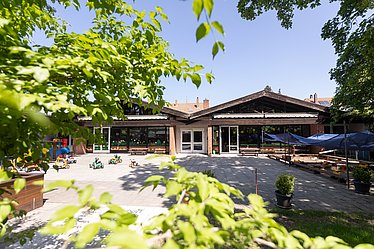 Es ist der Innenhof eines Kindergartens zu sehen, umzäunt von Bäumen. Auf der rechten Seite ist ein Sandkasten, links stehen einige Bobycars. Im Hintergrund ist die Einrichtung zu sehen.