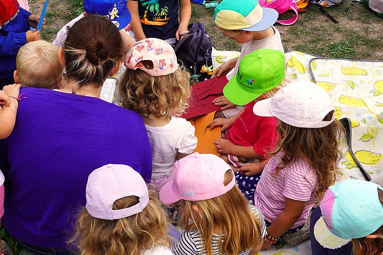 Es ist eine erwachsene Frau mit braunem Zopf zu sehen, die zusammen mit vielen Kindern auf einer bunten Decke sitzt. Die meisten der Kinder tragen eine Cappi
