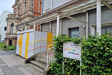 Es ist ein graues Gebäude zu sehen. Vor dem Gebäude geht eine kleine Treppe hoch. Rechts daneben steht in bunten Buchstaben Kinderkrippe Kuschelecke auf eine weißen Schild. Links neben der Treppe ist ein weiß-gelb gestreifter Container mit einer Sonne.