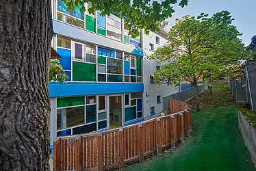 Links ist ein dicker Baumstamm zu sehen, daneben ein Gebäude mit blauen und grünen Scheiben, in Unterschiedlicher Größe, davor ein Gartenzaun.