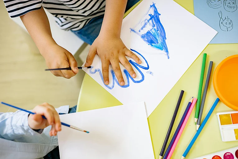 Es sind mehrere Kinderhände zu sehen, ein Kind mal einen Umriss seiner Hand mit einem blauen Stift auf ein weißes Papier