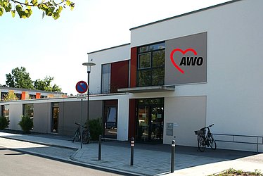 Es ist ein weißes Gebäude zu sehen, auf dem das Logo der AWO (ein halbes Herz und die Buchstaben AWO) angebracht ist.