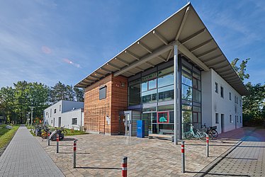 Es ist ein Gebäude zu sehen, der Eingangsbereich ist komplett verglast, links daneben hat das Gebäude eine Holz-Fassade, ansonsten ist es weiß. Der Himmel ist blau.