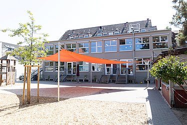 Es ist der Innenhof einer Kindertagesstätte zu sehen, mit einem großen Sandkasten, darüber ein orangenes Sonnensegel und im Hintergrund die Einrichtung, in grauer Fassade