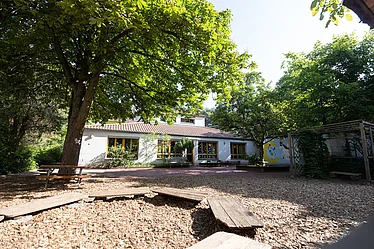 Es ist der Garten eines Kindergartens zu sehen, vorne ein sehr großer Sandkasten, nebendran große, grüne Bäume und im Hintergrund da Gebäude der Einrichtung.