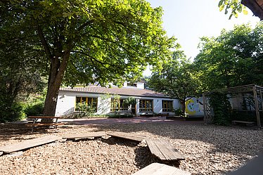 Es ist der Garten eines Kindergartens zu sehen, vorne ein sehr großer Sandkasten, nebendran große, grüne Bäume und im Hintergrund da Gebäude der Einrichtung.